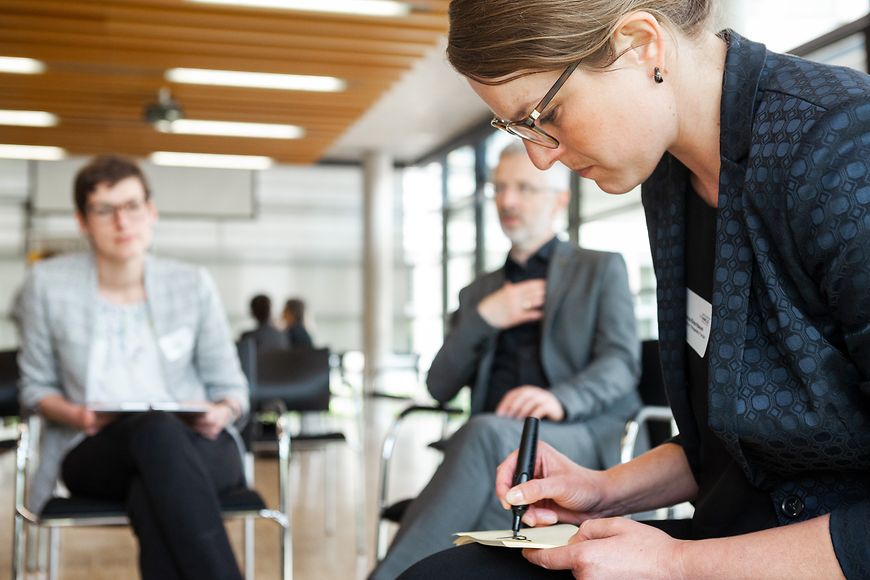 Eine Frau die mit einem Textmarker auf ein Zettel schreibt und zwei Personen die zuschauen